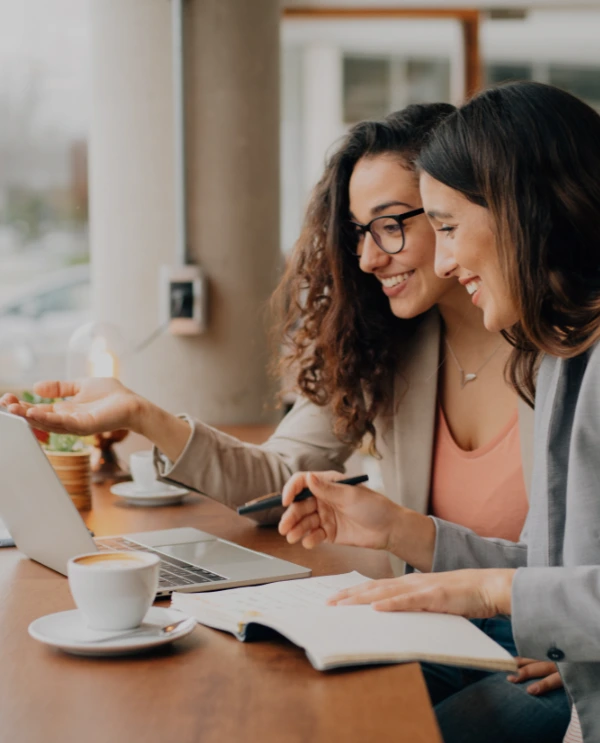 Dos mujeres empresarias sonrientes trabajando en una laptop en una cafetería, tomando notas y planificando juntas.