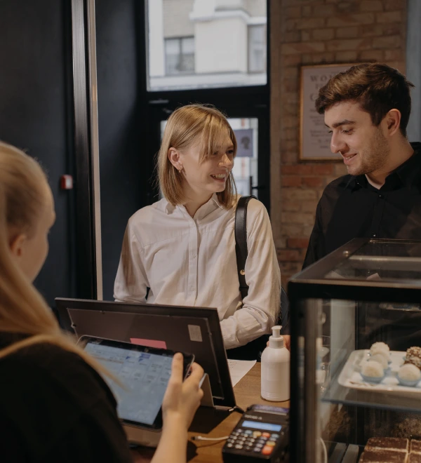 Pareja joven sonriendo mientras realiza un pedido en una cafetería con una empleada usando una tablet en la caja registradora.